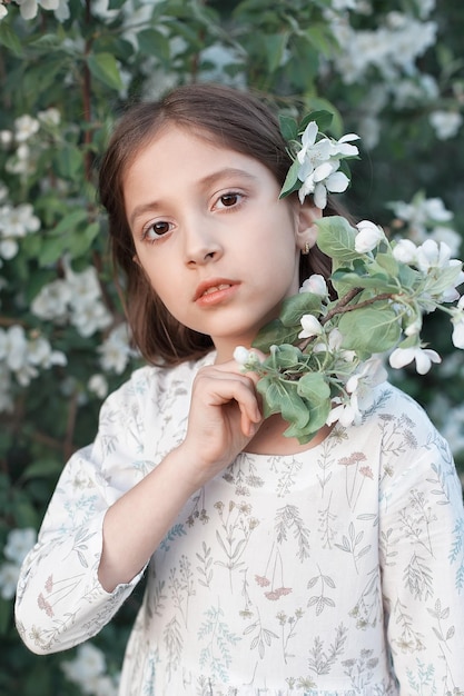 A beautiful little girl among the green foliage and white flowers of blooming apple trees Spring day A happy child smiles and looks at the camera