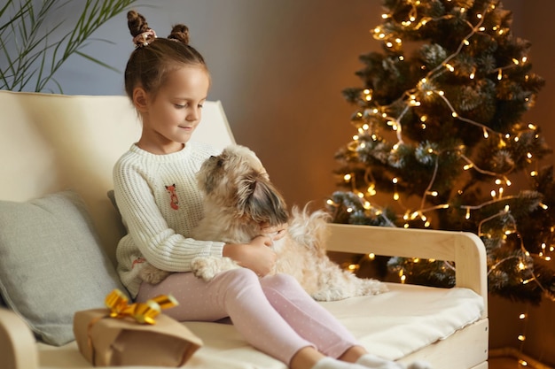 Beautiful little girl kid with funny hair buns playing with her favorite Pekingese dog on cough Girl cares about pet at home and celebrating Christmas together