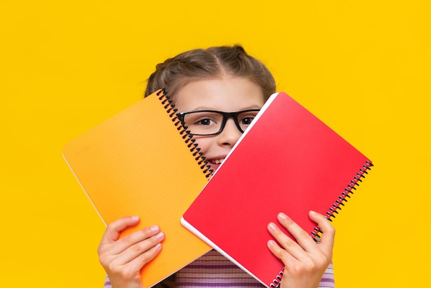 A beautiful little girl looks out from behind a red notebook and smiles A schoolgirl on a yellow isolated background