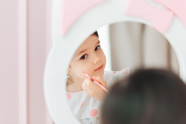 A beautiful little girl in the mirror preening a little girl is sitting at a children's table and is wearing children's cosmetics