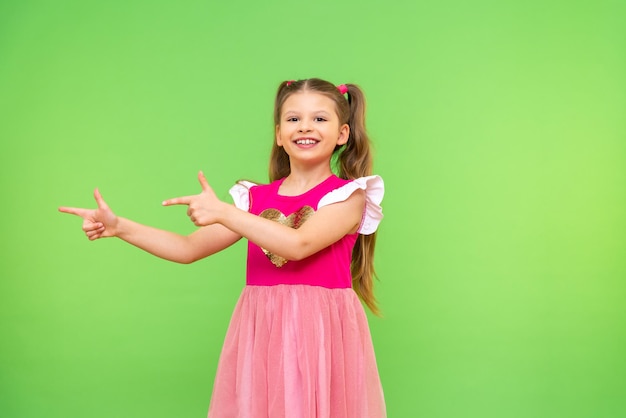 A beautiful little girl in a pink dress points with her hands at the advertisement Children on a green isolated background