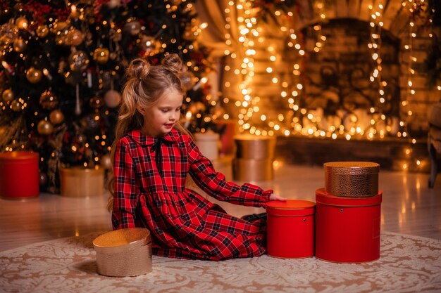 Beautiful little girl in a red dress at home near the tree opens gifts