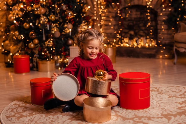 Beautiful little girl in a red dress at home near the tree opens gifts