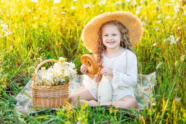 Beautiful little girl sitting in a straw hat in a yellow field with wild flowers with a bottle of milk and a bagel