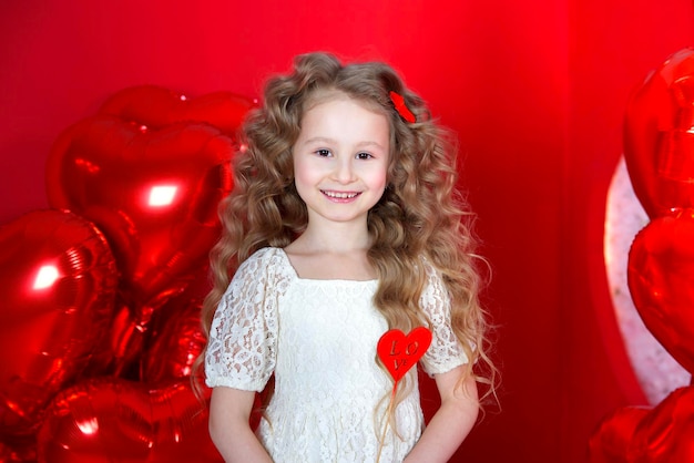 Beautiful little girl smiles on a red background with a heartshaped balloon and a gift box in her hands The concept of love gifts Valentine39s Day February 14 March 8 mother39s day