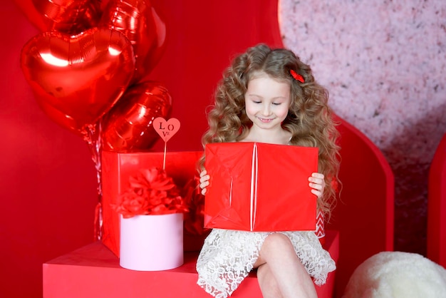 Beautiful little girl smiles on a red background with a heartshaped balloon and a gift box in her hands The concept of love gifts Valentine39s Day February 14 March 8 mother39s day