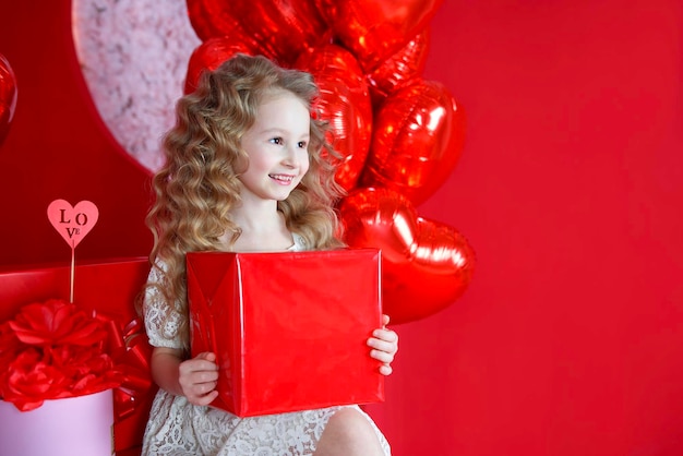 Beautiful little girl smiles on a red background with a heartshaped balloon and a gift box in her hands The concept of love gifts Valentine39s Day February 14 March 8 mother39s day