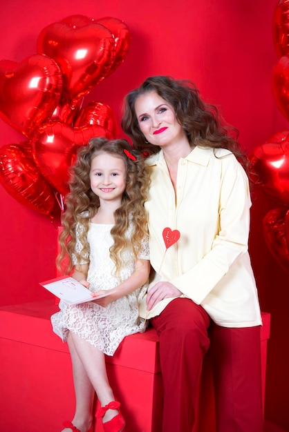 Beautiful little girl smiles on red background with heartshaped balloon and a gift box in her hands The concept of love present Valentine39s Day February 14 March 8 mother39s day vertical photo