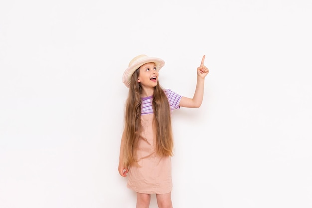 A beautiful little girl in a summer sundress and a straw hat on a white isolated background points her index finger up at your advertisement