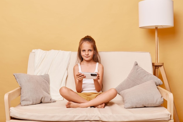 Beautiful little girl wearing casual clothing sitting on sofa at home against beige wall holding mobile phone looking at camera enjoying playing video games watching cartoons on phone