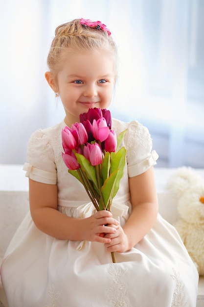 Beautiful little girl in a white dress sitting on a windowsill with a bouquet of tulips