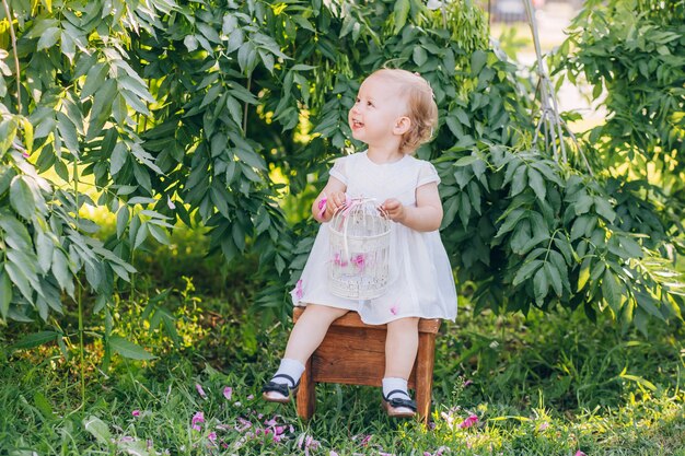 Beautiful little girl with blond hair in a white dress sitting on a chair in the park sunny summer day.