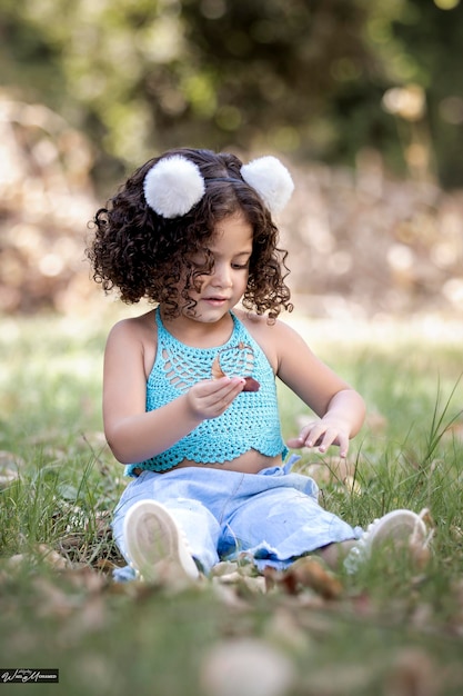 Beautiful little girl with curly hair sitting on the grass holding fallen leaves while having fun