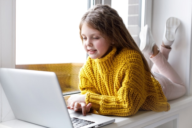 A beautiful little girl with a laptop at home at the window looks at the screen and smiles