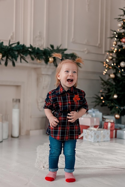 Beautiful little girl with ponytails stands near the Christmas tree with gifts and laughs