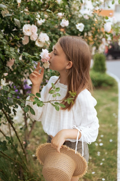 Beautiful little girl with a straw hat near a bush of white roses