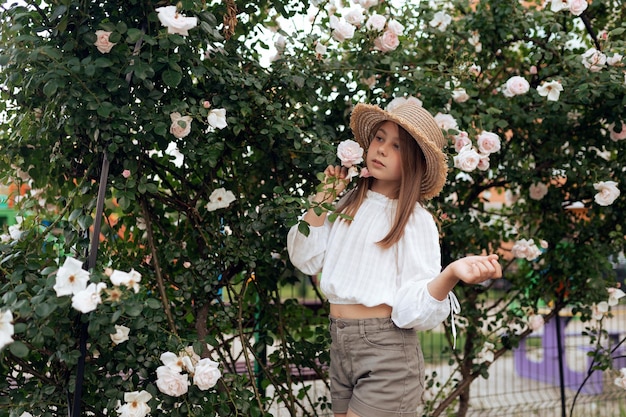 Beautiful little girl with a straw hat near a bush of white roses