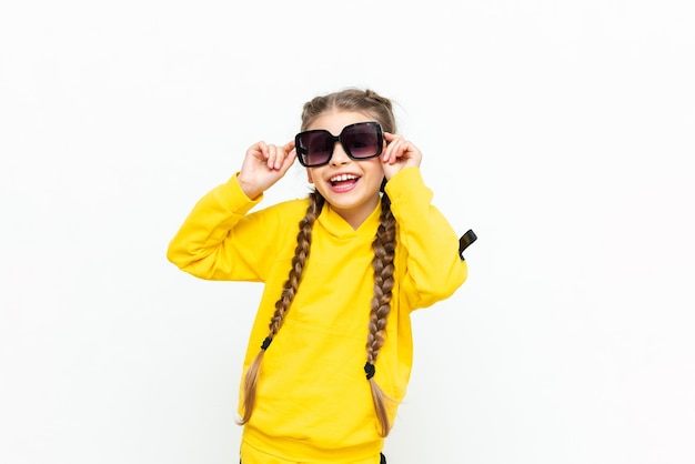 A beautiful little girl in a yellow tracksuit with pigtails on her head and sunglasses on a white isolated background