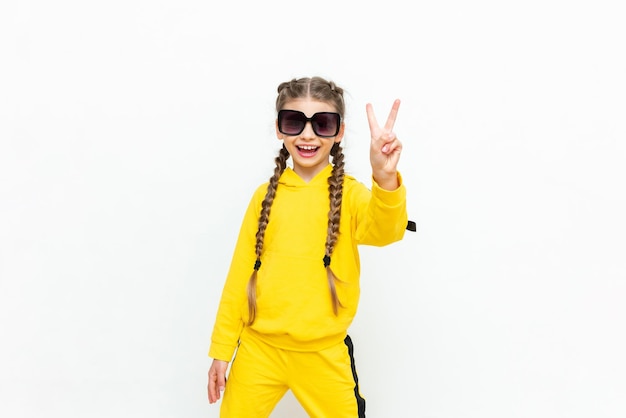 A beautiful little girl in a yellow tracksuit with pigtails on her head and sunglasses on a white isolated background