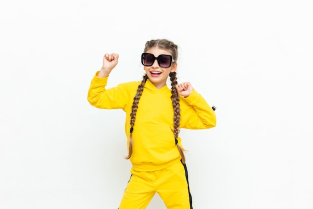 A beautiful little girl in a yellow tracksuit with pigtails on her head and sunglasses on a white isolated background