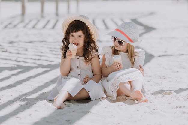 Beautiful little girls in white dress and hat eating ice cream on beach in summer time