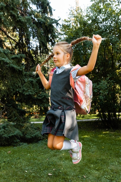 A beautiful little schoolgirl with a pink backpack is walking in the park and having fun. School uniform.