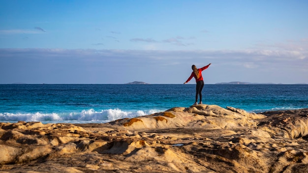 Photo beautiful long-haired girl stands on rocks above the ocean with her hands raised, enjoying freedom