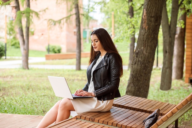 Beautiful long haired woman, student is working, communicating, studying using her laptop outdoors in park in summer.