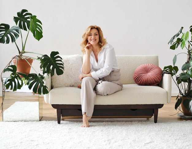 Beautiful mature blonde woman sits behind on the sofa looks into the camera and smiles There are pots with live monsters in the room