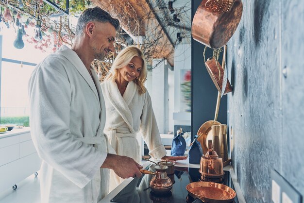 Beautiful mature couple in bathrobes preparing coffee together while spending time in the domestic k...