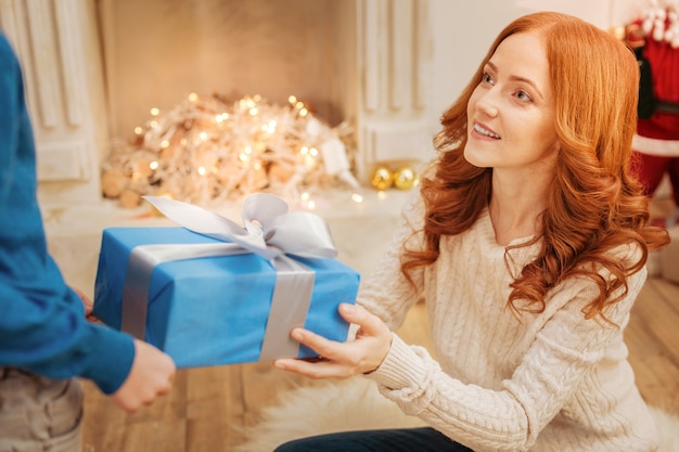 beautiful mature lady sitting on the floor while giving her little son a Christmas present on a festive morning in a family circle.