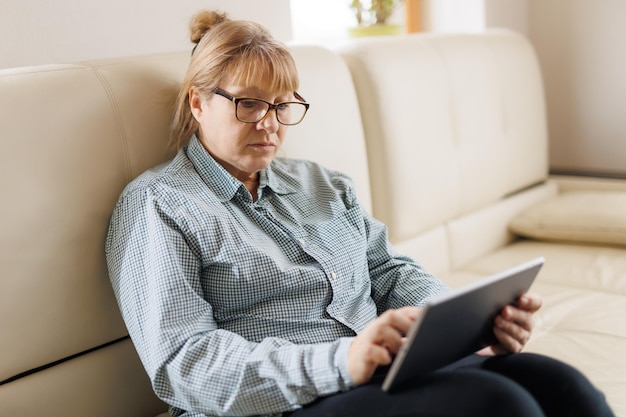 Beautiful mature woman in eyeglasses is using a digital tablet and smiling while sitting on couch at home