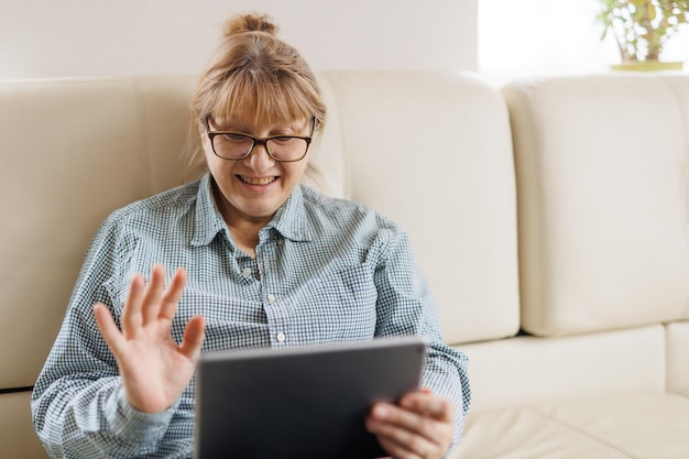 Beautiful mature woman in eyeglasses is using a digital tablet and smiling while sitting on couch at home