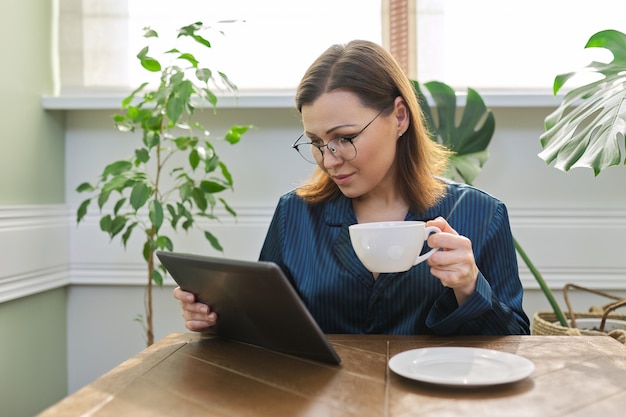 Beautiful mature woman in pajamas reading news, social networks, mail in digital tablet. Morning portrait of middle-aged female at home at table with cup of tea, breakfast.