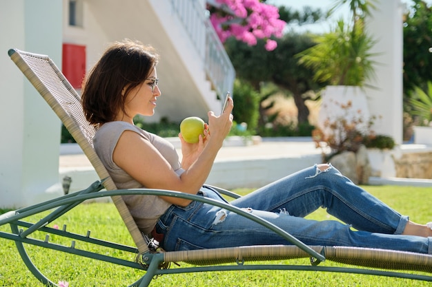 Beautiful mature woman resting on sunbed on green lawn. Relaxing female eating apple and using smartphone, sunny summer day. Lifestyle, leisure, nutrition, middle-aged people