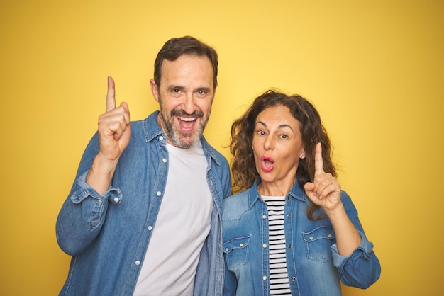 Beautiful middle age couple together wearing denim shirt over isolated yellow background pointing finger up with successful idea Exited and happy Number one