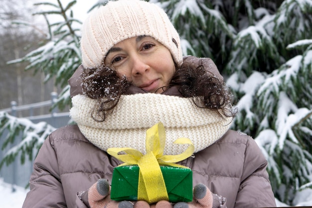 A beautiful middle-aged 45-year-old woman holding a nice Christmas present in the air. Snowy landscape with spruce and snow