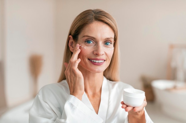 Beautiful middle aged woman in silky bathrobe using moisturizing eye cream enjoying face care routine