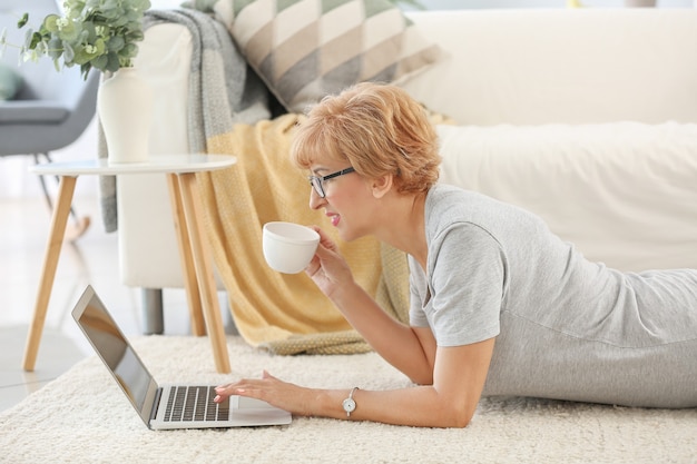 Beautiful middle-aged woman with laptop and cup of tea resting at home