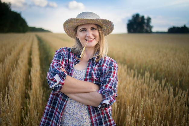 A beautiful middleaged farmer woman in a straw hat and a plaid shirt stands in a field of golden ripening wheat during the daytime in the sunlight