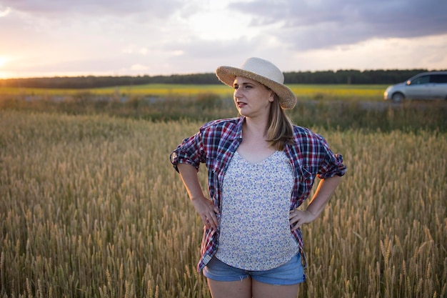 A beautiful middleaged farmer woman in a straw hat and a plaid shirt stands in a field of golden ripening wheat during the daytime in the sunlight