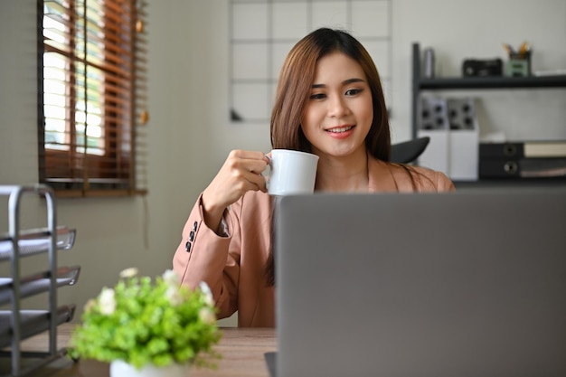 Beautiful millennial Asian businesswoman looking at laptop screen while sipping morning coffee