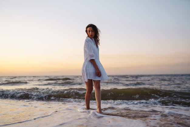 Beautiful model in elegant white dress at sunset with glass of wine The girl looks at the sunrise