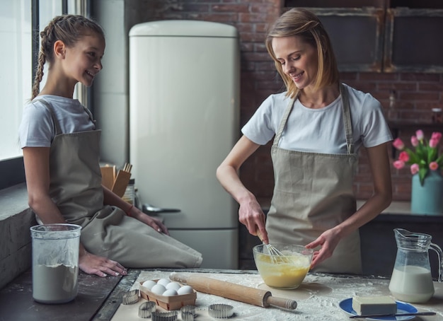 Beautiful mom and daughter in aprons are making cookies at home mom is whisking eggs