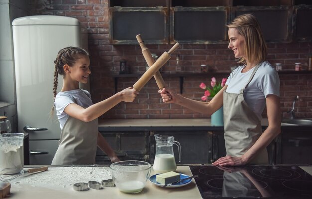 Beautiful mom and daughter in aprons are pretending to fight with rolling pins looking at camera and smiling while cooking at home