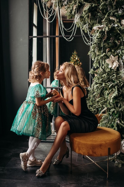 Beautiful mom and daughter in stylish holiday dresses for Christmas near the Christmas tree Mom and adorable daughter pose near the Christmas decorations New Year Christmas