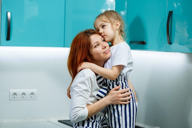 Beautiful mother and daughter are hugging and smiling joyfully, sitting in the kitchen. Family, children, motherhood and cooking concept. Happy family relationships