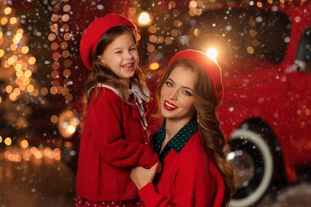 Beautiful mother and daughter in red dresses on the background of Christmas lights and red retro car