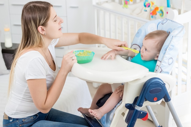 Beautiful mother feeding her baby boy in highchair with porridge