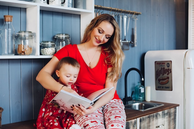 Beautiful mother with little daughter in Christmas pajamas reading book at kitchen.